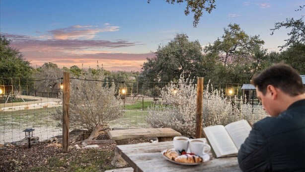 A wooden picnic table with a book, cups, and pastries, set against a backdrop of trees and a sunset. - Charming Barndominium with Farm Animals – Just 5 Minutes from Lake Georgetown (Georgetown)