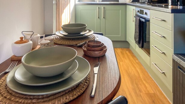 A kitchen with light green cabinets, a wooden table with plates and bowls, and a microwave on the counter. - Ddwylig Bach - UK48623 (Rhyl and Prestatyn)