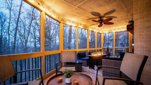 Un patio techado con una chimenea, mobiliario de mimbre y ventanas amplias con vista a árboles.