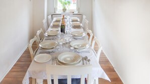 Una larga mesa de comedor preparada para una comida, con sillas blancas, platos, cubiertos y una botella de champán.