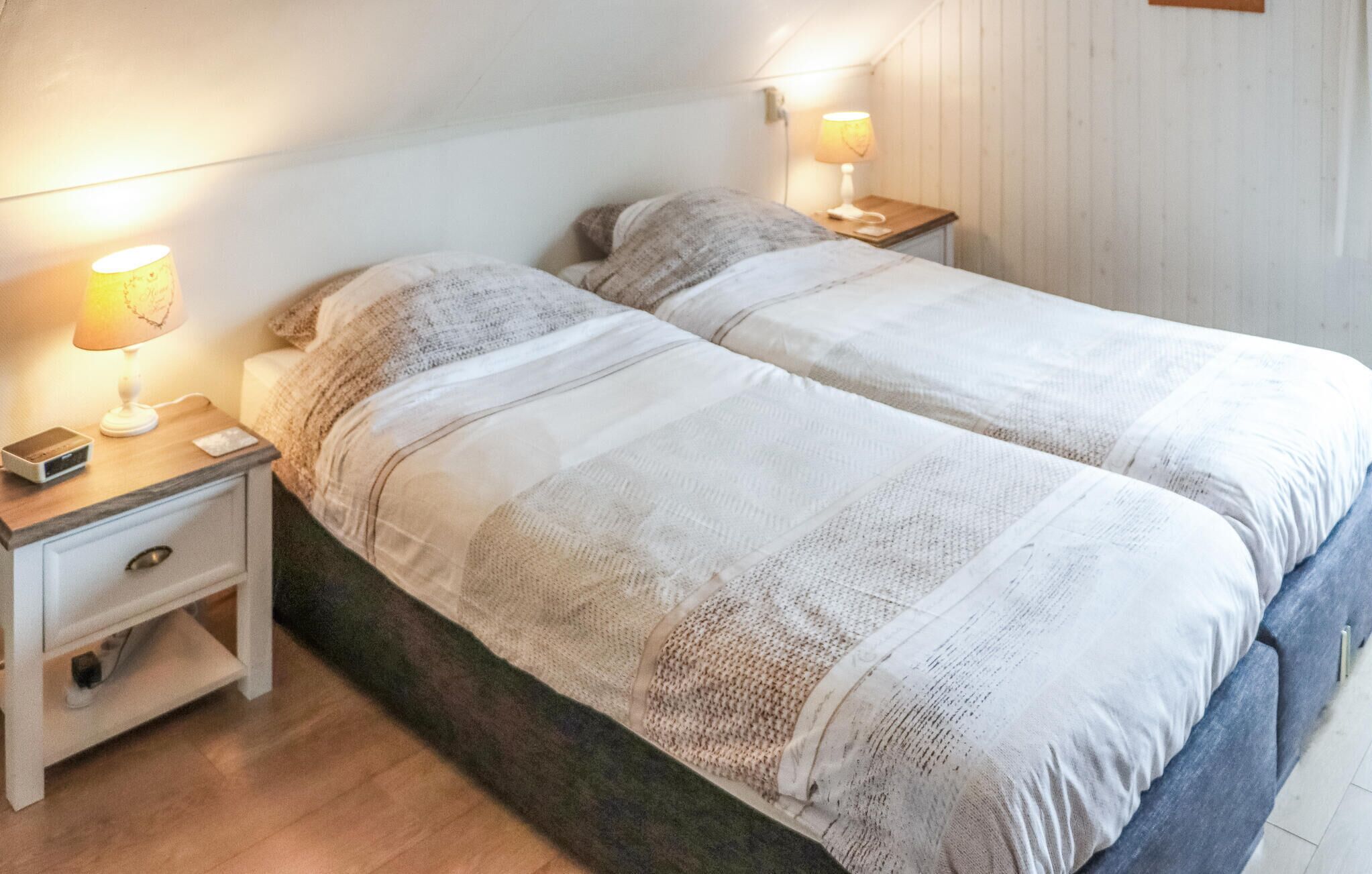 Ein Doppelbett mit einer weiß-grauen Streifenoptik, zwei Nachttischlampen, ein Nachttischchen mit einem Radio und ein Holzboden.