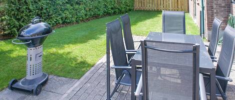 Een terras met een tafel, stoelen, een barbecue en een haag op de achtergrond.