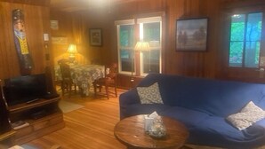 A cozy room with a blue sofa, a wooden coffee table, a TV, and a dining area with a table and chairs. - Angola on the Lake Beach Front House  4BR  private beach wrap-around porches (Angola)