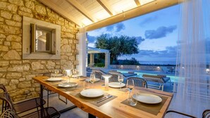 An outdoor dining area with stone walls, a wooden table set for six, and a view of the sea and trees. - Ionian Stone Luxury Villa Aether (Corfu)