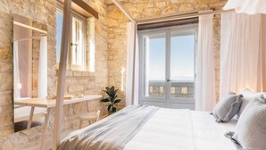 A bedroom with a stone wall, a large bed with grey bedding, a desk, and a view of the sea through a large window. - Ionian Stone Luxury Villa Aether (Corfu)