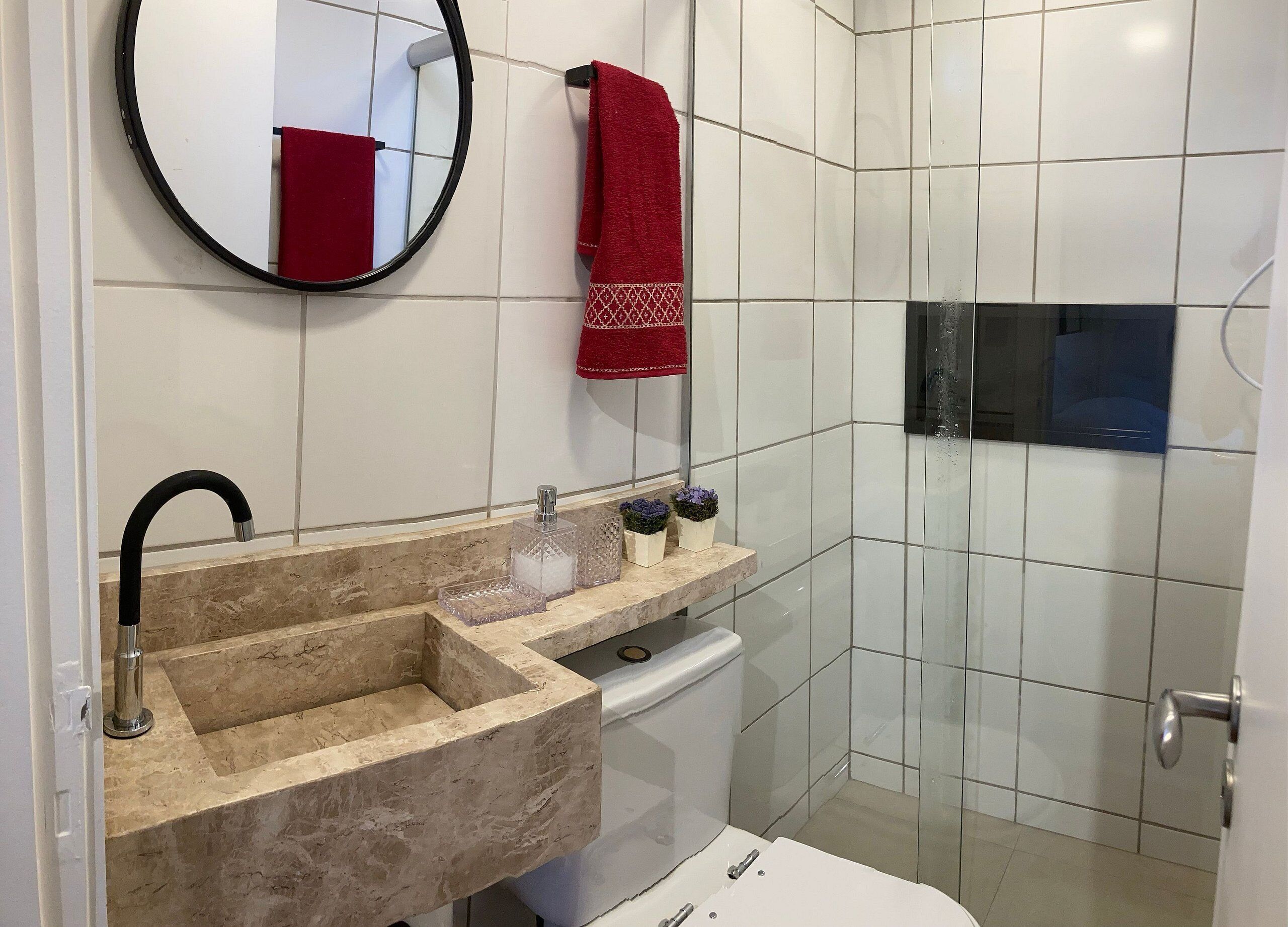 A bathroom with a beige marble sink, a round mirror, a red towel, and a glass shower enclosure.