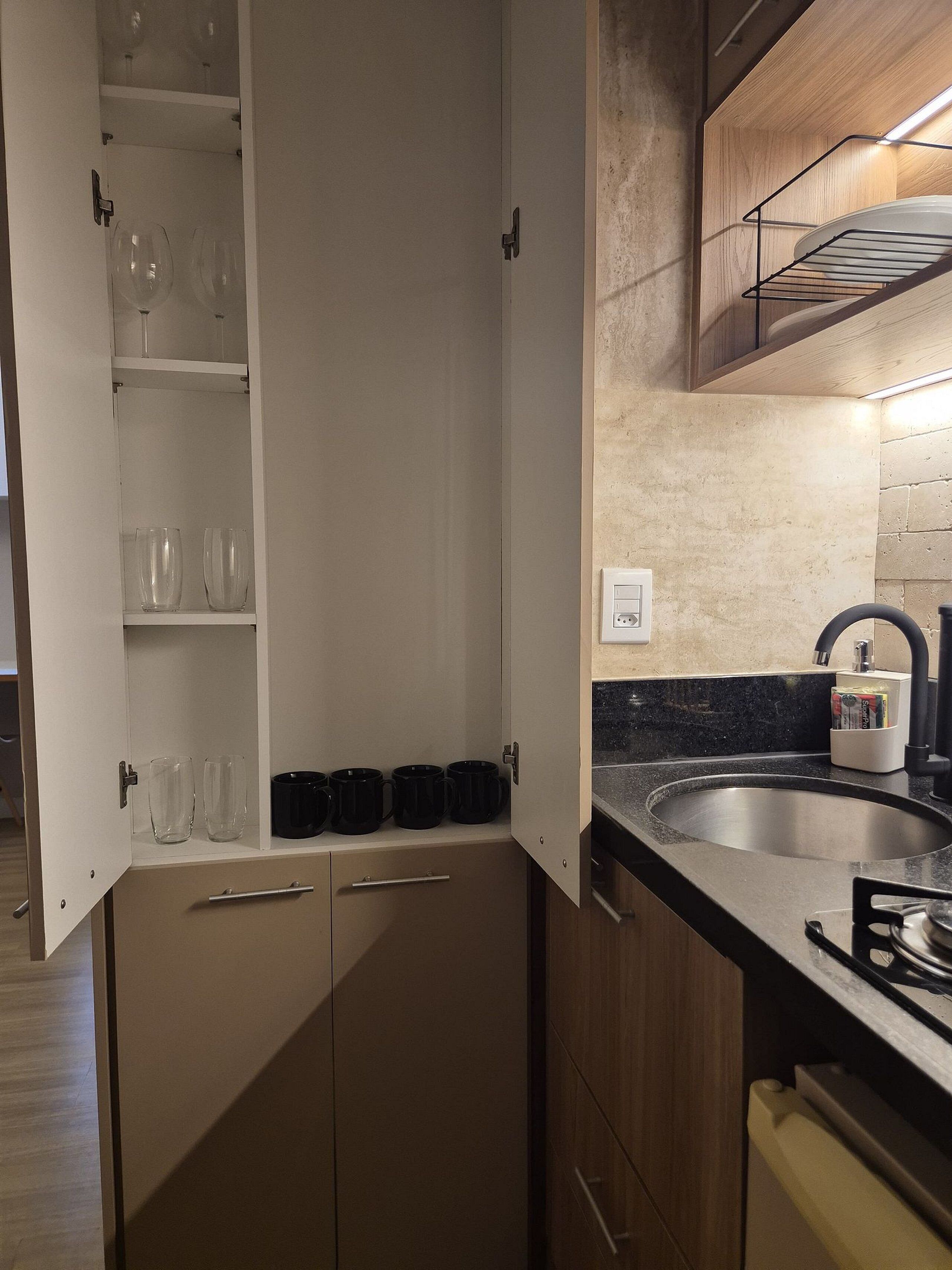 Een compacte keuken met een inbouwoven, een spoelbak en een kast met drinkglazen en zwarte mokken.