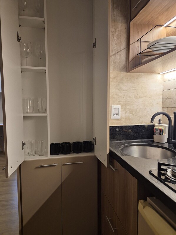 A compact kitchen with a built-in oven, a sink, and a cabinet with glassware and black mugs.