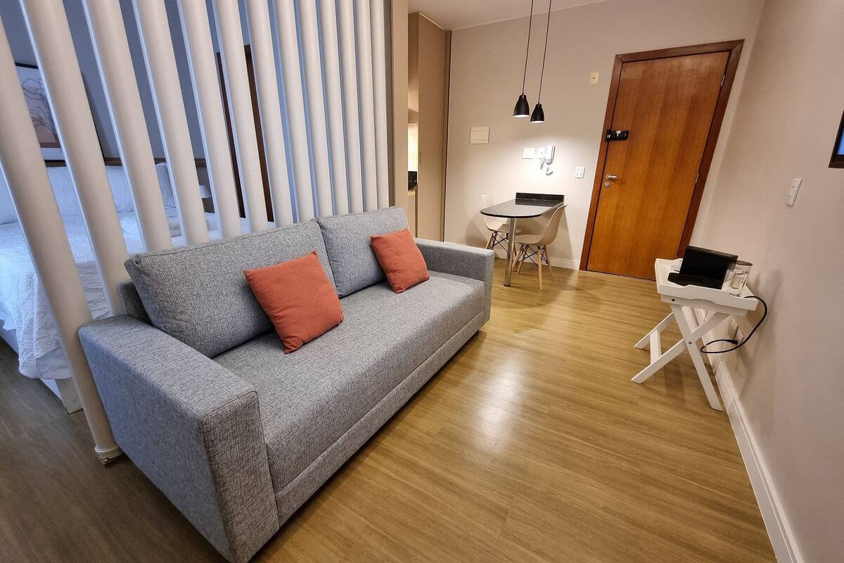 Una sala de estar moderna con un sofá gris, dos cojines naranjas, piso de madera, una mesita blanca con una computadora portátil y un comedor con una mesa pequeña y sillas.