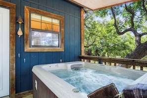 A hot tub with a wooden deck, a window with a view of trees, and a wooden door. - Stronghold Retreat | 3 Cabins by Creek w/ Hot Tubs (Bandera)
