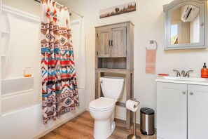 A bathroom with a shower curtain, toilet, sink, cabinet, and a trash can. - Stronghold Retreat | 3 Cabins by Creek w/ Hot Tubs (Bandera)