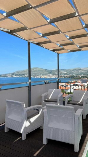 A covered outdoor seating area with wicker chairs, overlooking a coastal town and the sea.