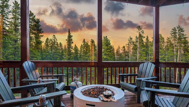 A wooden deck with Adirondack chairs, a fire pit, and a scenic view of a forest and sunset. - Sauna• Hot tub• 2 King Beds• Game Room• Mtn views• Close to Breck• Pet friendly (Fairplay)