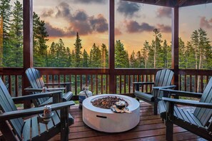 A wooden deck with Adirondack chairs, a fire pit, and a scenic view of a forest and sunset. - Luxe• Mtn views• Sauna• Hot tub• 2 King Beds• Game Room• Close to Breck (Fairplay)