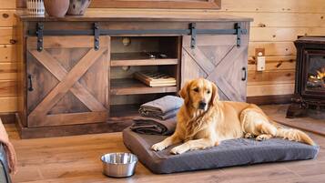 Een hond rustend op een comfortabel bed in een kamer met houten wanden, een open haard en een metalen kom op de vloer.