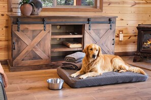 Un perro descansando sobre una cama acolchada en una habitación con paredes de madera, una chimenea y un tazón de metal en el suelo.