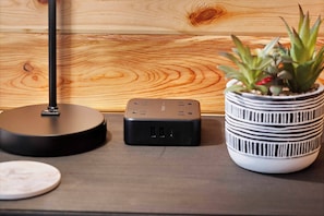 Una mesa moderna con un altavoz negro, un dispositivo pequeño negro, y una planta en maceta con hojas verdes y flores rojas.