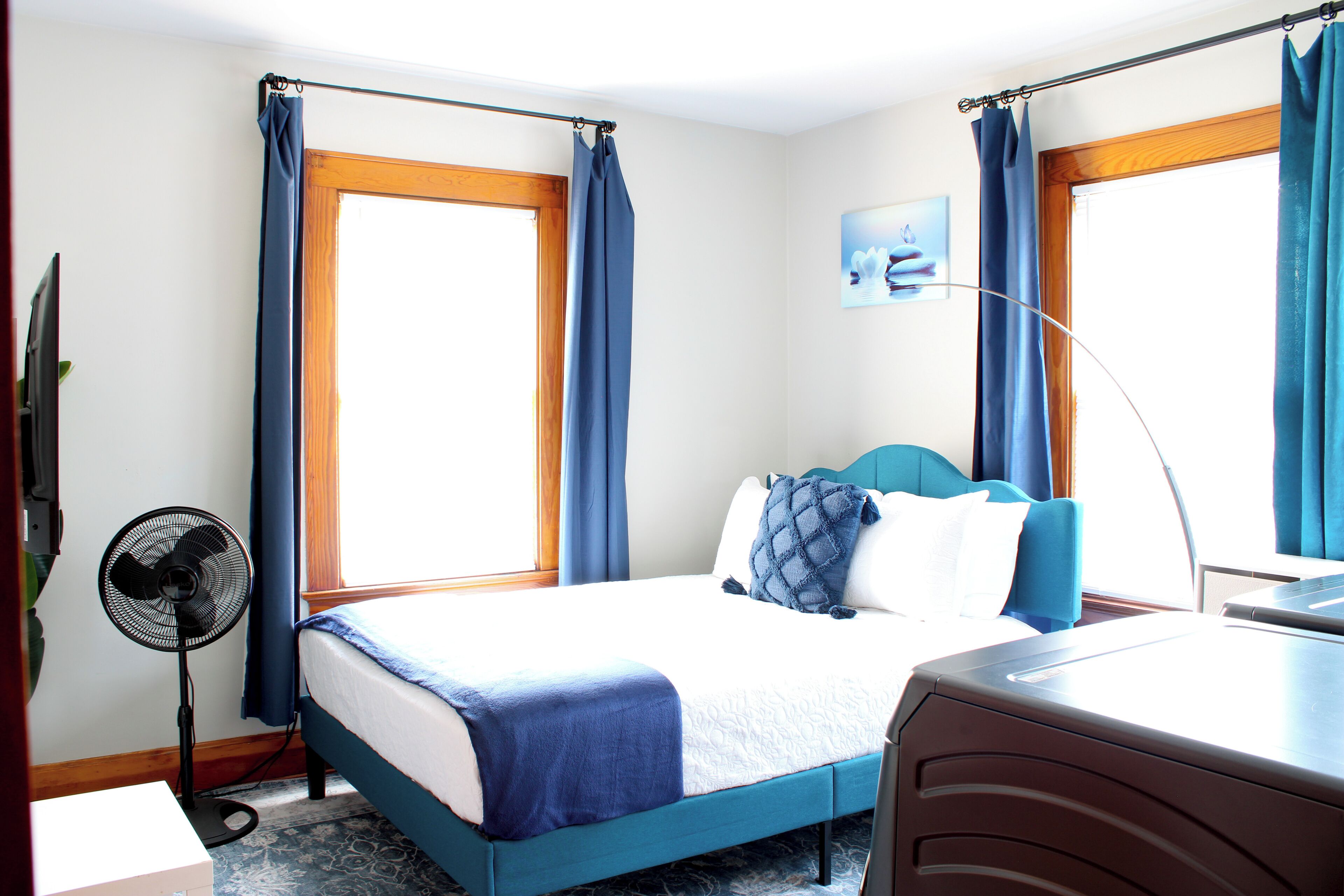 Una habitación de hotel con una cama, un escritorio, una silla, un ventilador de pie y dos ventanas con cortinas.