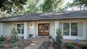 A single-story house with a white brick exterior, a front porch with potted plants, and a wooden front door. - The Game Palace @ Sugar Magnolia (Jackson)
