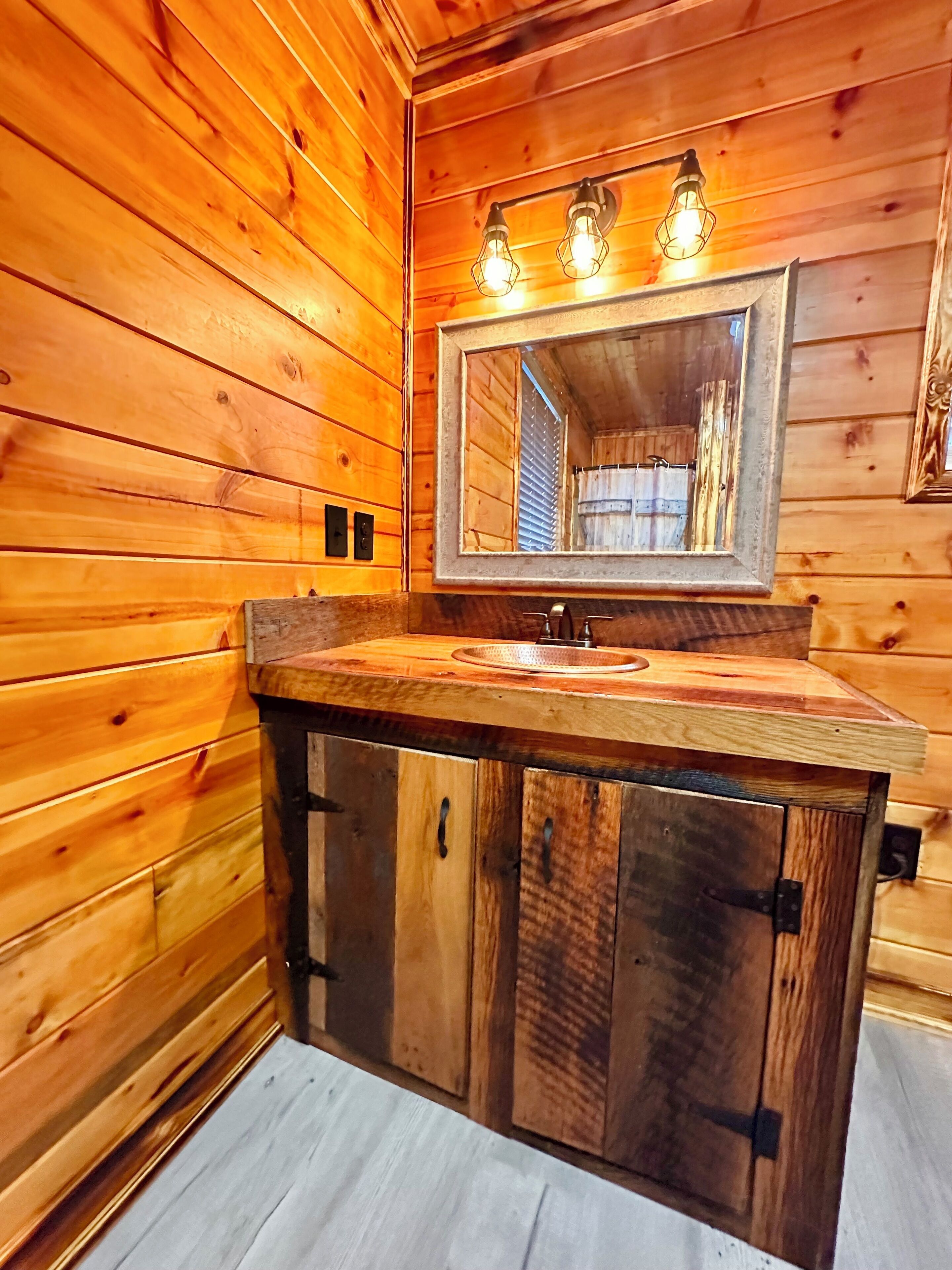 Baño de cabaña de madera con tocador de madera, un espejo y tres lámparas fijadas a la pared.
