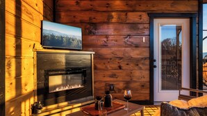 Ein gemütliches Blockhaushaus-Interieur mit Kamin, einem darüber montierten Fernseher und einem Tisch mit einem Weinglas und einer Kerze.
