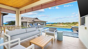A spacious outdoor area with a pool, seating, and a view of a house and ocean. - Amaui Island Escape New 4-bedroom home! Free Access to Mauna Kea & Hapuna Beach! (Kamuela)
