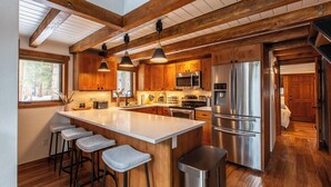 A modern kitchen with wooden beams, a large island, bar stools, stainless steel appliances, and a view of the outdoors. - A-Frame Cabin with Private Sauna & Hot Tub (Breckenridge)