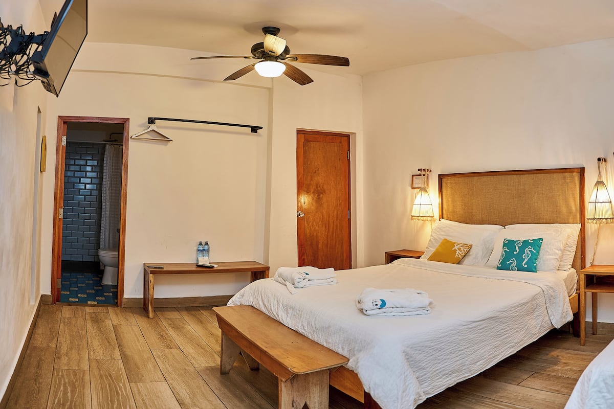 A hotel room with a wooden floor, a bed with white bedding, a wooden bench, a bedside table with a lamp, a ceiling fan, and a bathroom visible through an open door.