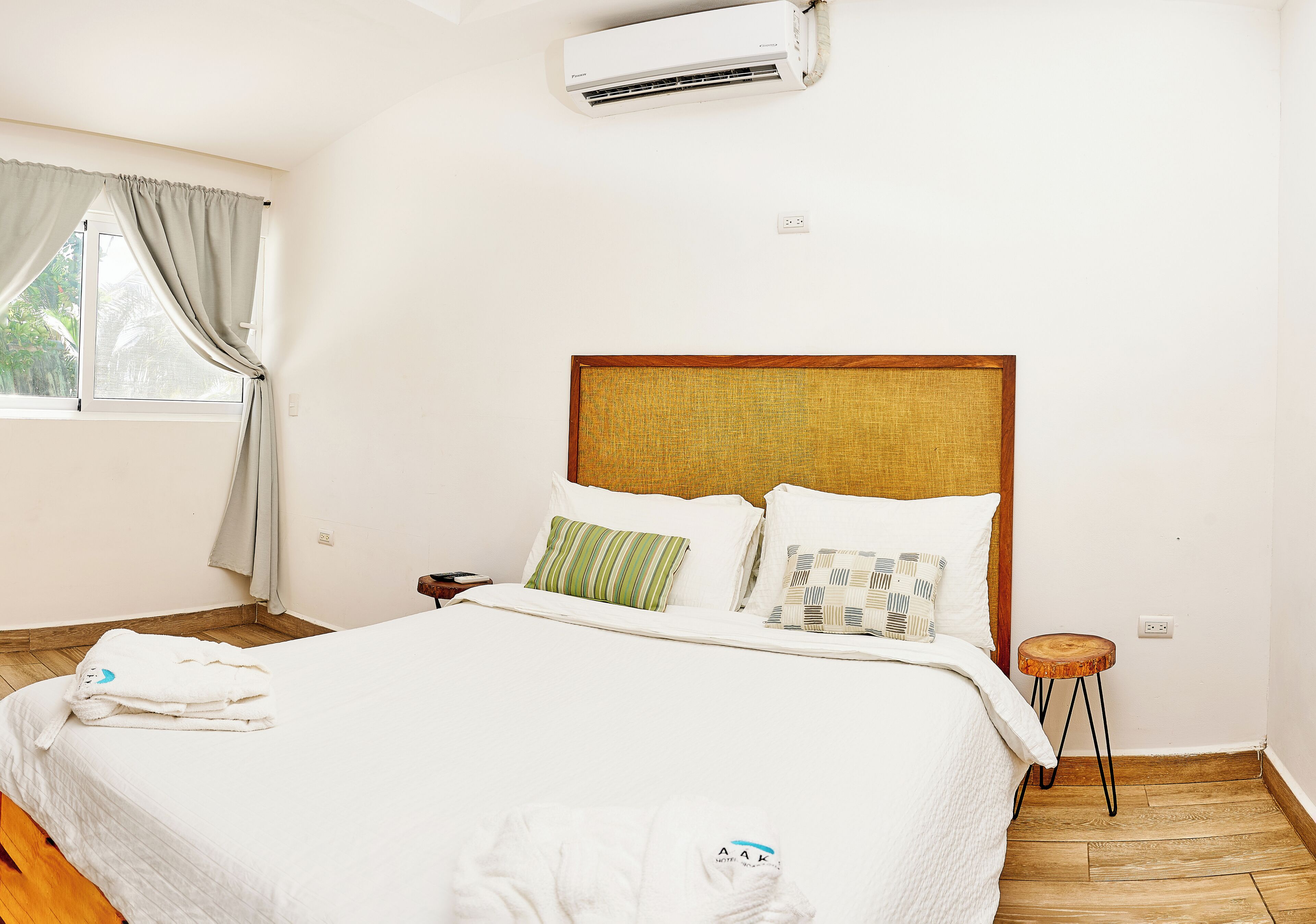 Habitación de hotel con cama, mesita de noche, piso de madera, ventana con cortinas y equipo de aire acondicionado.