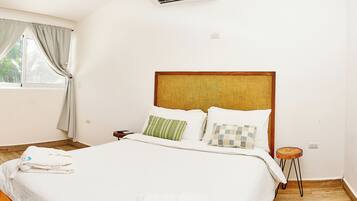 Habitación de hotel con cama, mesita de noche, piso de madera, ventana con cortinas y equipo de aire acondicionado.