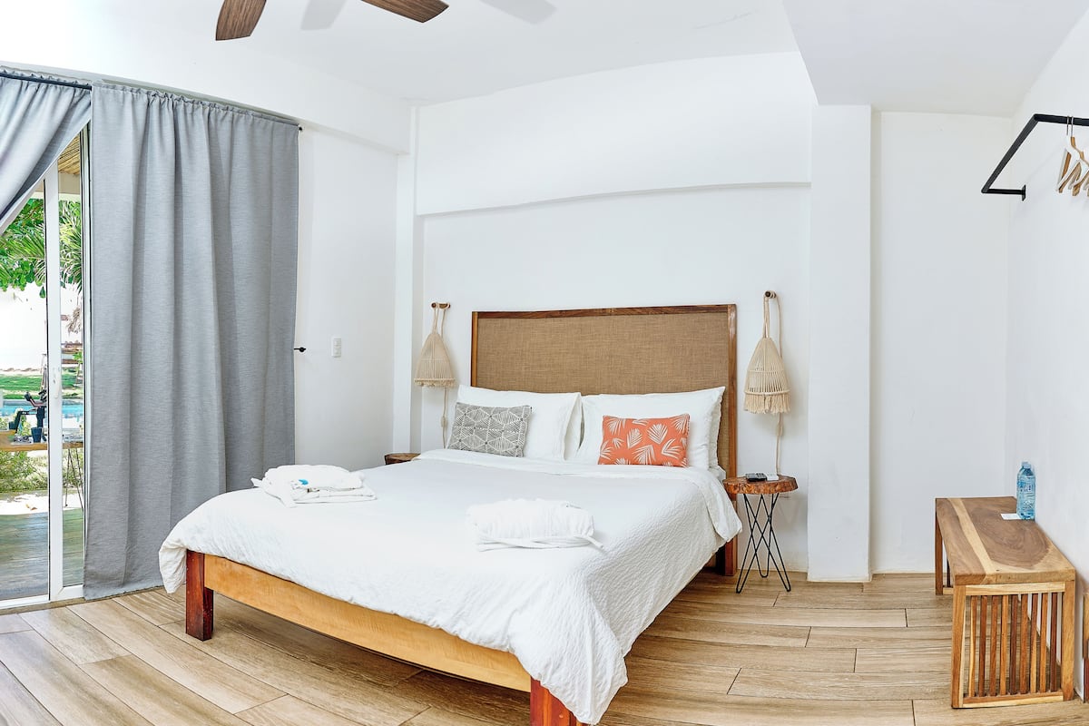 A modern bedroom with a wooden bed, a ceiling fan, a bedside table with a lamp, and a view of the outdoors.