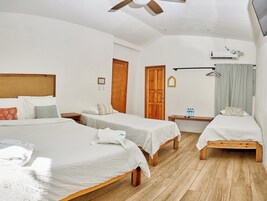 Habitación de hotel con dos camas individuales, cabecero de madera, ventilador de techo, unidad de aire acondicionado, mesita de noche con una botella de agua y un banco de madera.