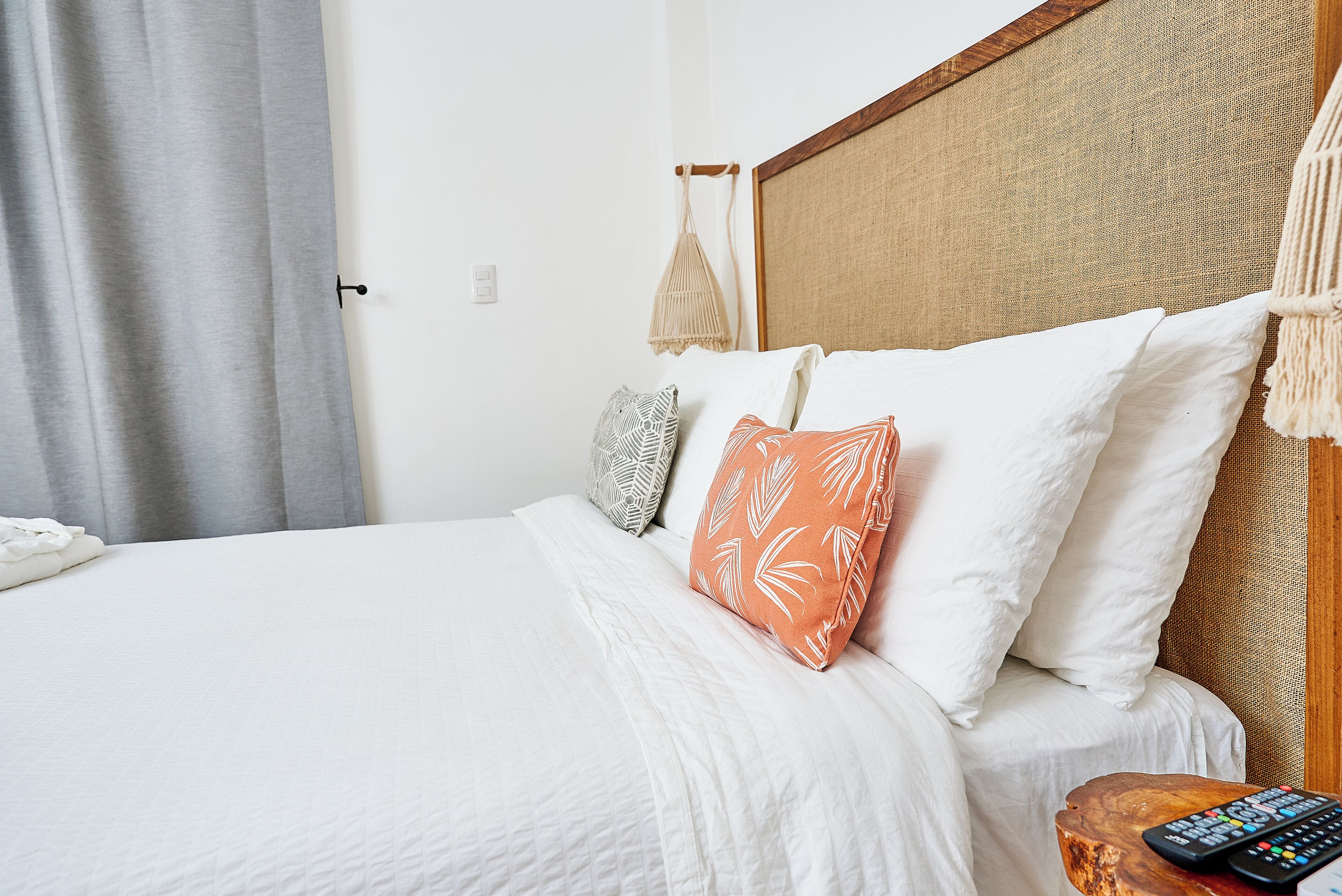 A neatly made bed with white bedding and patterned pillows, a wooden headboard, and a remote control on a wooden side table.