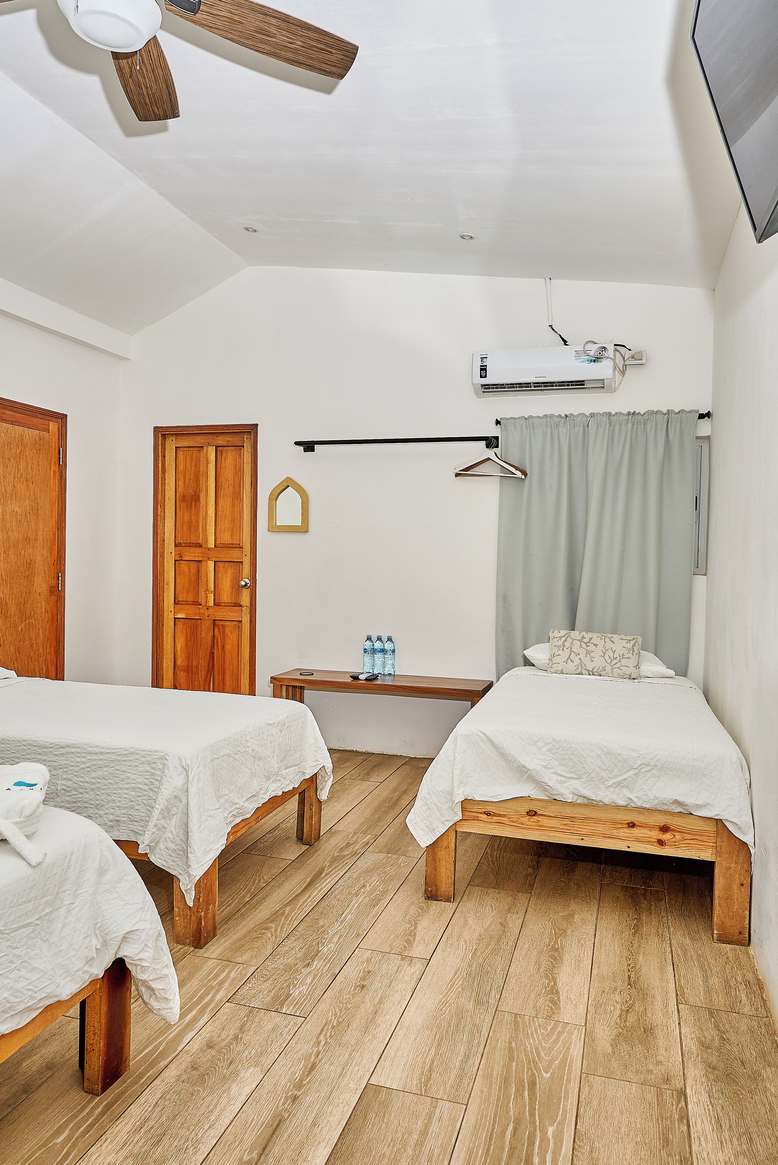 A room with two wooden beds, a ceiling fan, an air conditioning unit, a wooden bench, a small table with water bottles, and a door.
