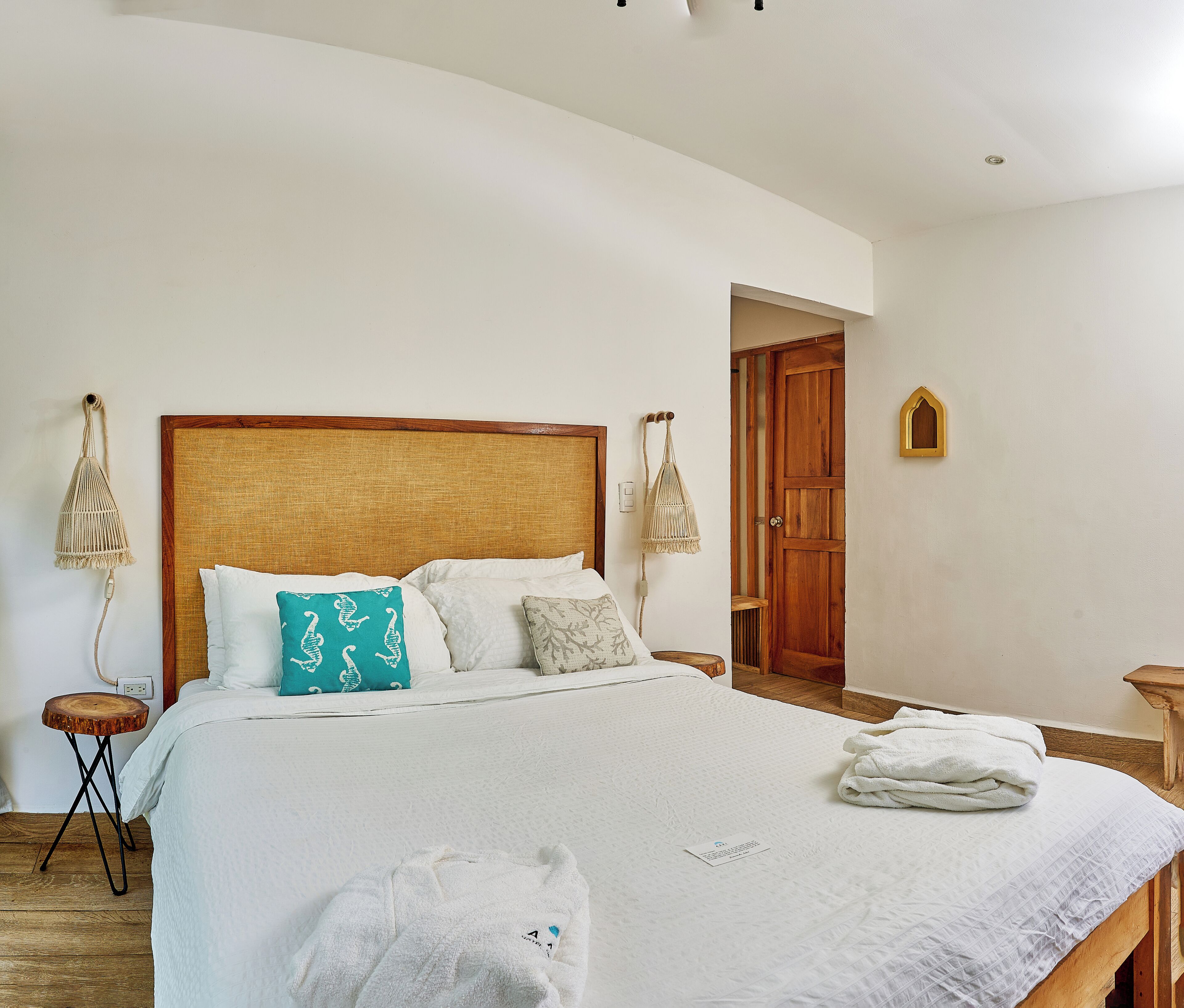 Una cama bien tendida con ropa blanca y cabecera de madera, una puerta de madera y una pequeña estantería en la pared.