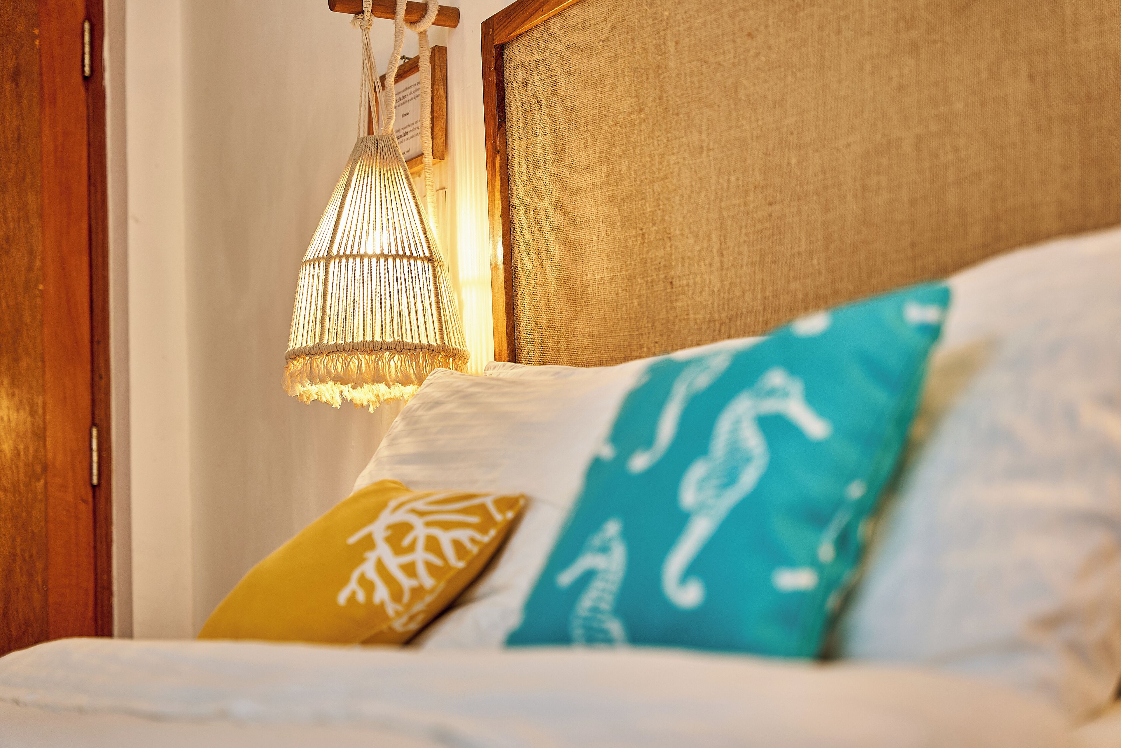 A bed with white bedding and two decorative pillows, one yellow and one blue, with a hanging light fixture above.