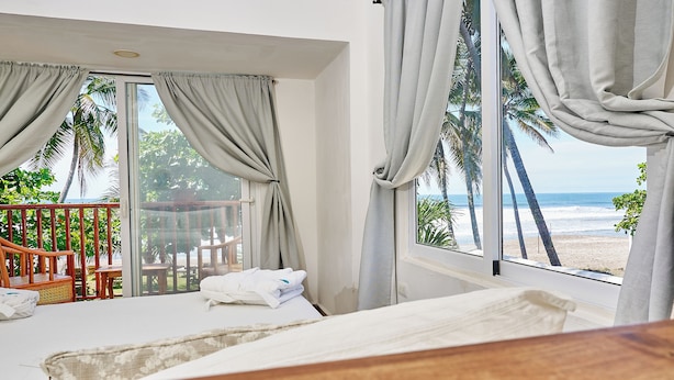 A hotel room with a bed, a wooden headboard, a balcony with a wooden chair and table, and a view of the beach and palm trees. - Aaki Hotel (León)