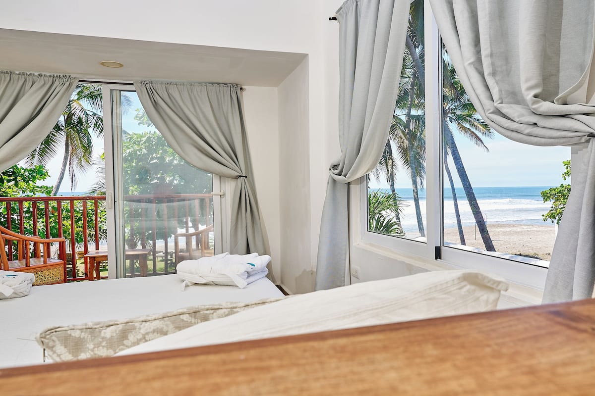 A hotel room with a bed, a wooden headboard, a balcony with a wooden chair and table, and a view of the beach and palm trees.