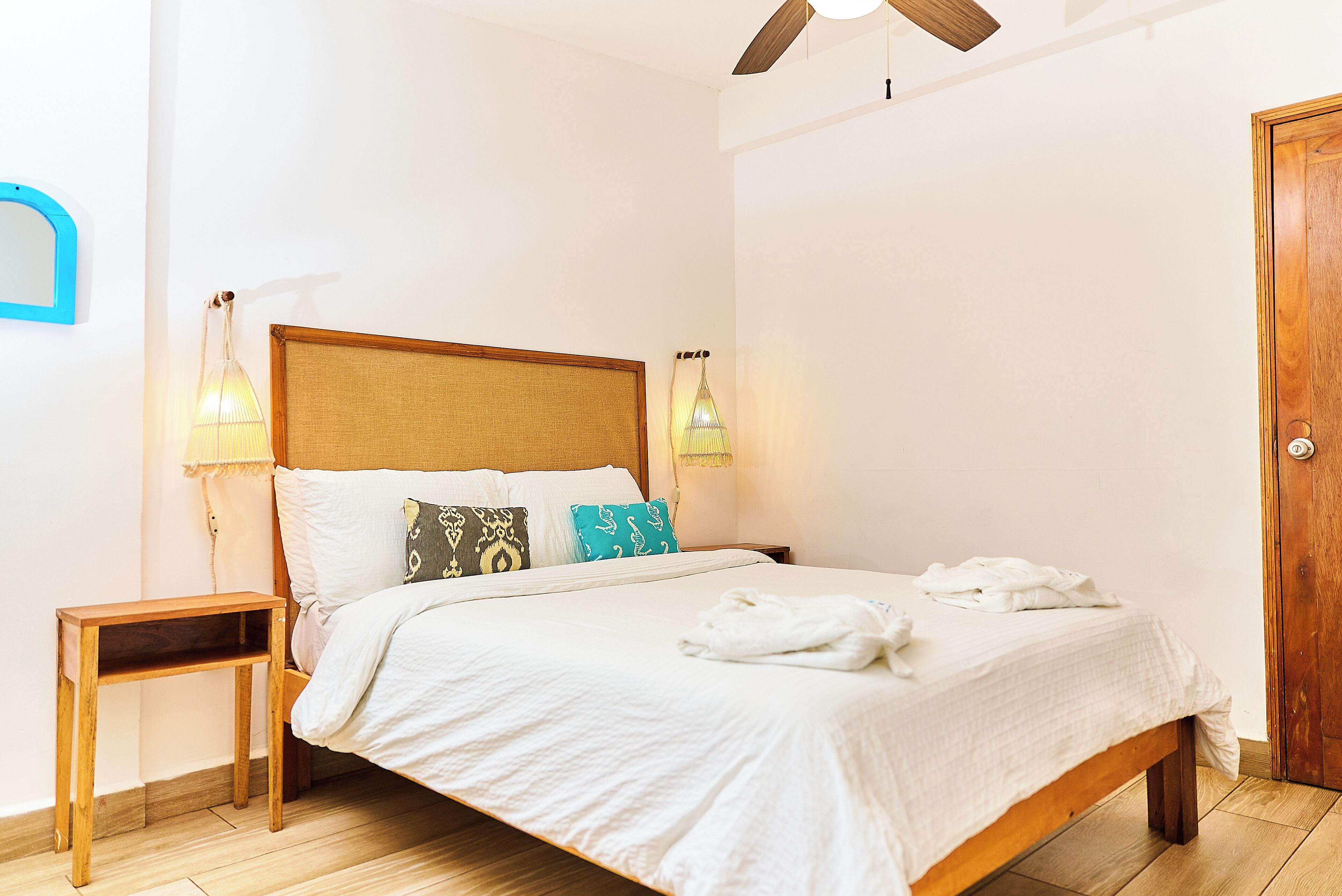 Un lit bien fait, avec du linge de lit blanc et plusieurs oreillers, une table de chevet en bois, deux lampes fixées au mur et une porte en bois.