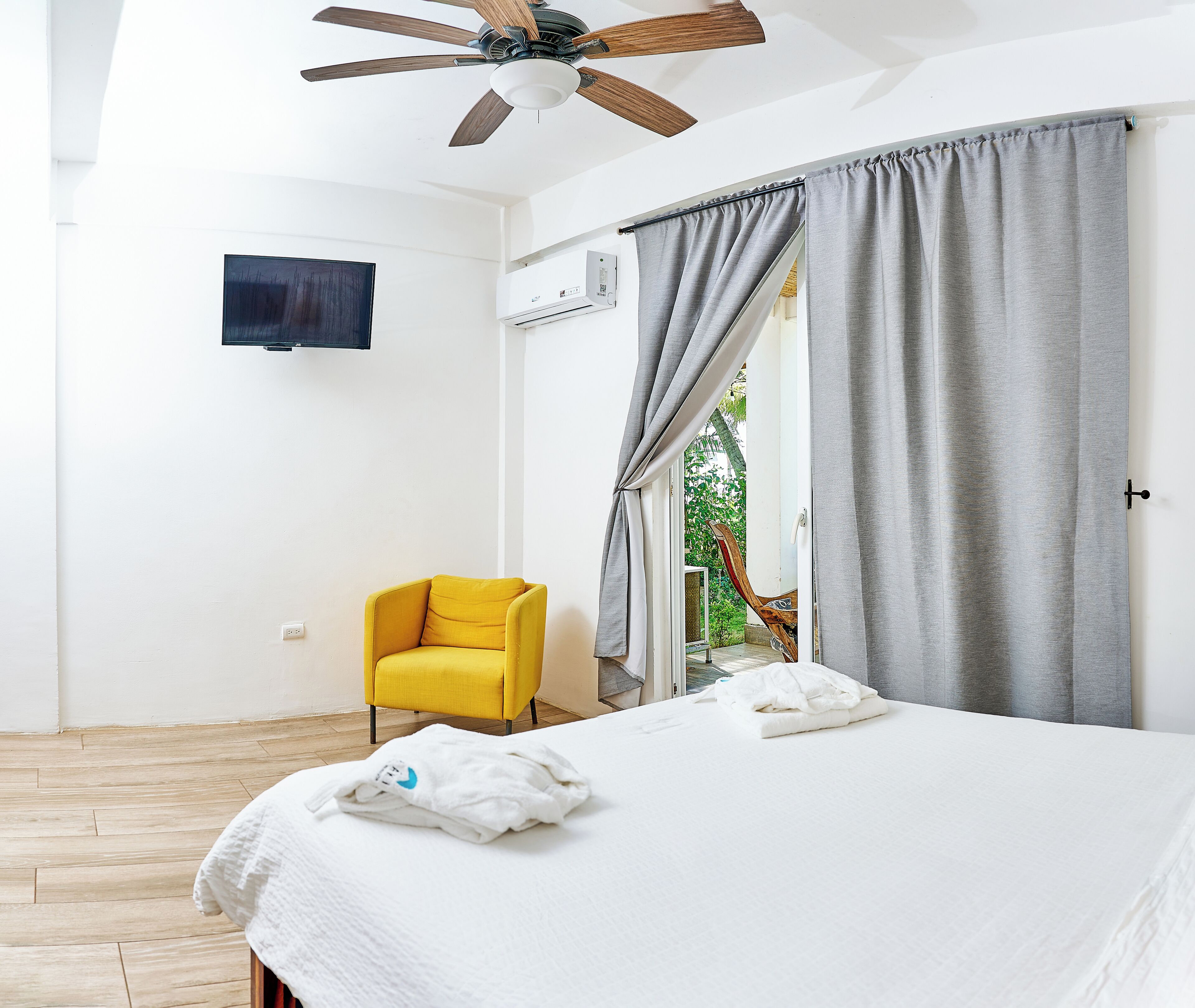 A bedroom with a bed, a yellow chair, a ceiling fan, and a window with grey curtains.