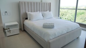 A modern bedroom with a large bed, a nightstand, and a view of greenery outside. - Apartamento en Baia Kristal (Cartagena)
