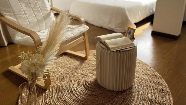 Een gezellige kamer met een houten stoel, een klein rond voetenbankje met een boek, en een bed met een witte dekbedovertrek.