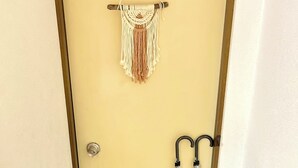 Entree van een hotelkamer met een houten deur, wanddecoratie van macramé, twee paraplu's en een paar slippers op de vloer.