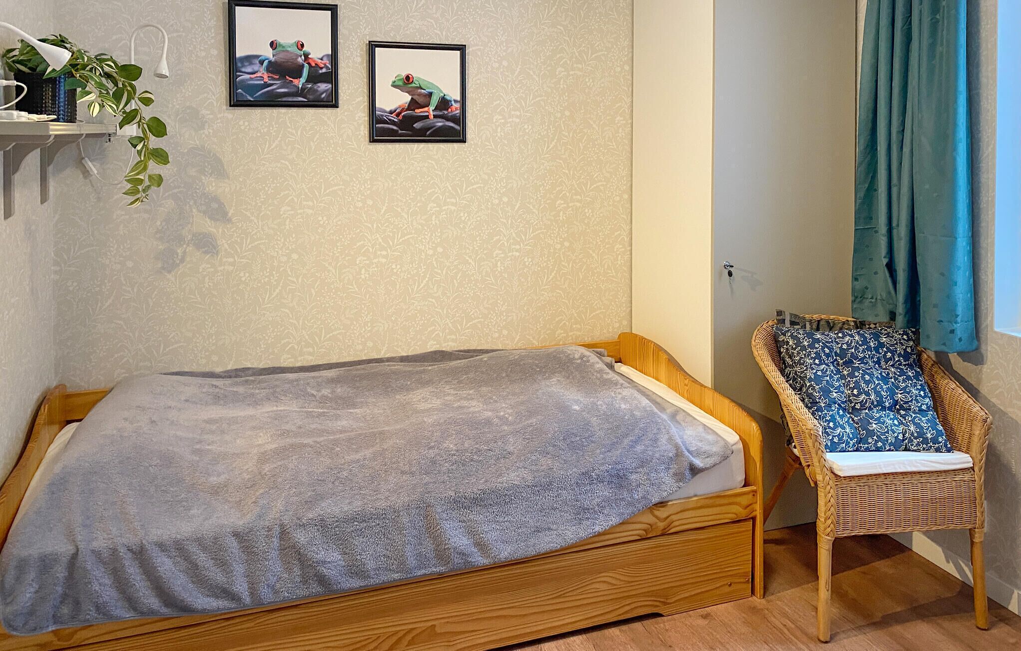 Uma cama de solteiro com um cobertor cinzento, uma cadeira de vime com uma almofada estampada e duas molduras na parede.