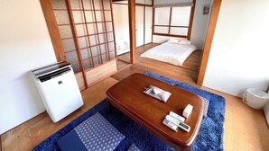 Um quarto tradicional japonês com tatames, um painel shoji, uma cama com roupa de cama branca, uma pequena mesa com uma toalha e um controle remoto, e um aparelho de ar condicionado branco.