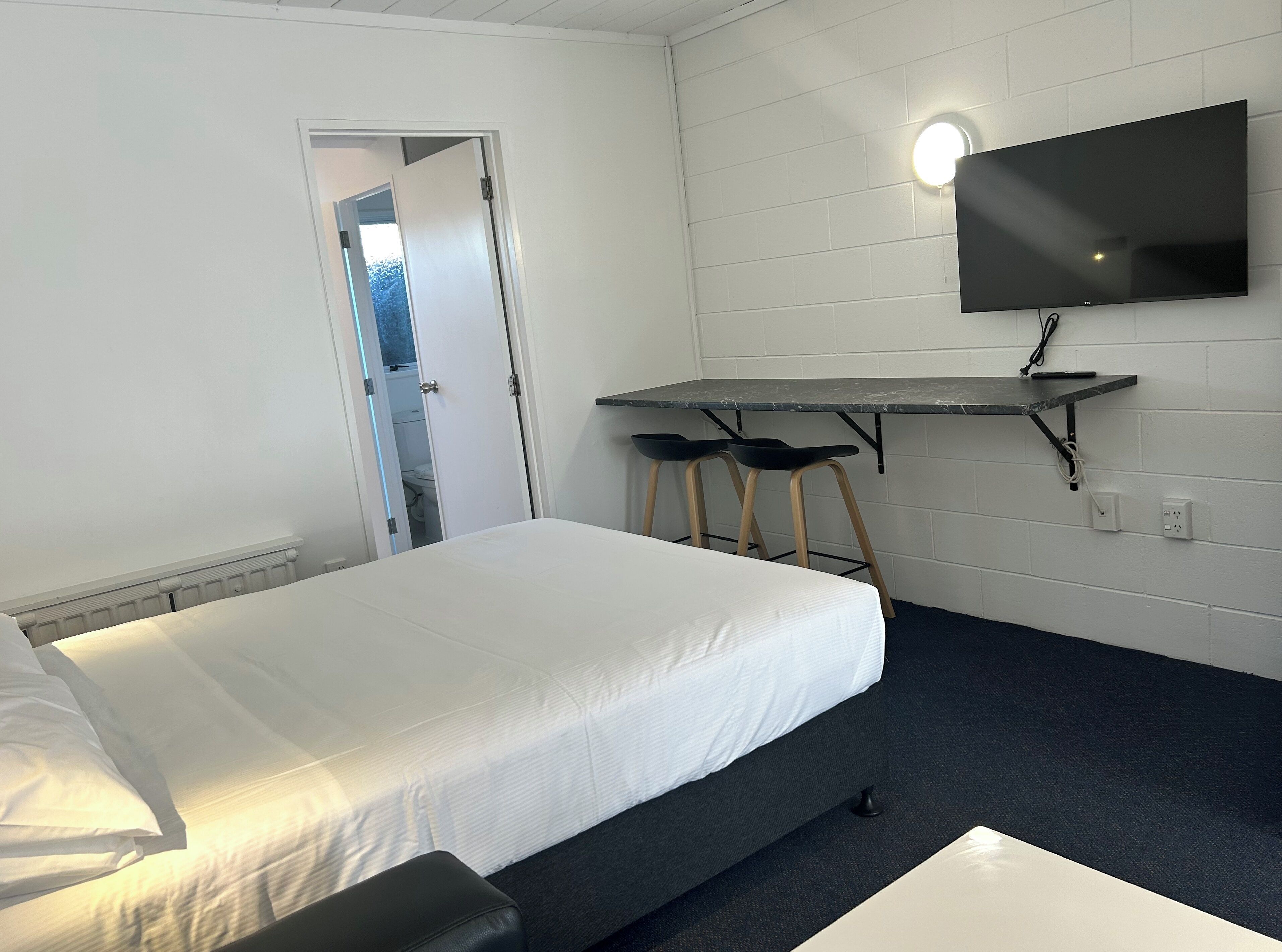 A room with a bed, a desk with two chairs, a TV mounted on the wall, and a door leading to another room.