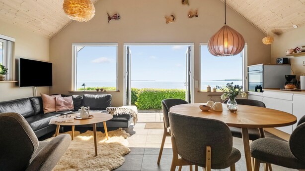 A modern living room with a black leather sofa, a round wooden coffee table, and a dining area with a wooden table and chairs. There are large windows offering a view of the sea. - 1 bedroom gorgeous home in Sønder Stenderup (Sønder Stenderup)