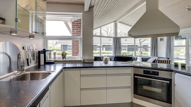 A modern kitchen with a stainless steel sink, white cabinets, a built-in oven, and large windows offering views of a residential area.