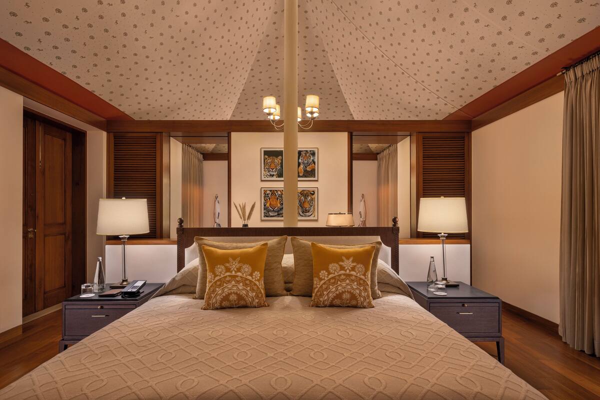 Quarto de hotel com uma cama grande, duas mesas de cabeceira, um criado-mudo, um lustre e um espelho amplo.
