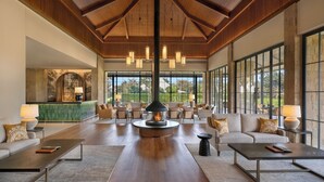 A spacious lobby with a high ceiling, large windows, and a central fireplace. Furnishings include sofas, armchairs, and coffee tables.